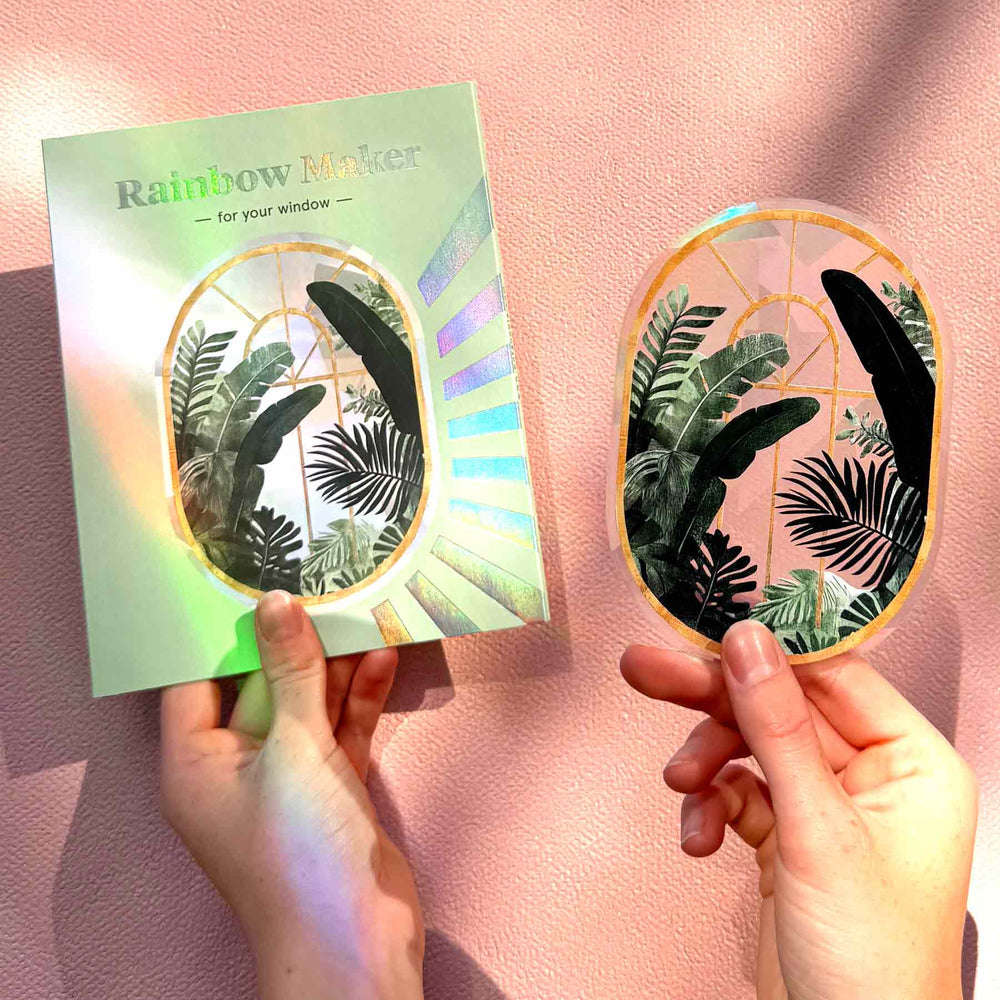 Hände halten den botanischen Fenstersticker und seine Rainbow-Maker-Verpackung vor einer rosafarbenen Wand.