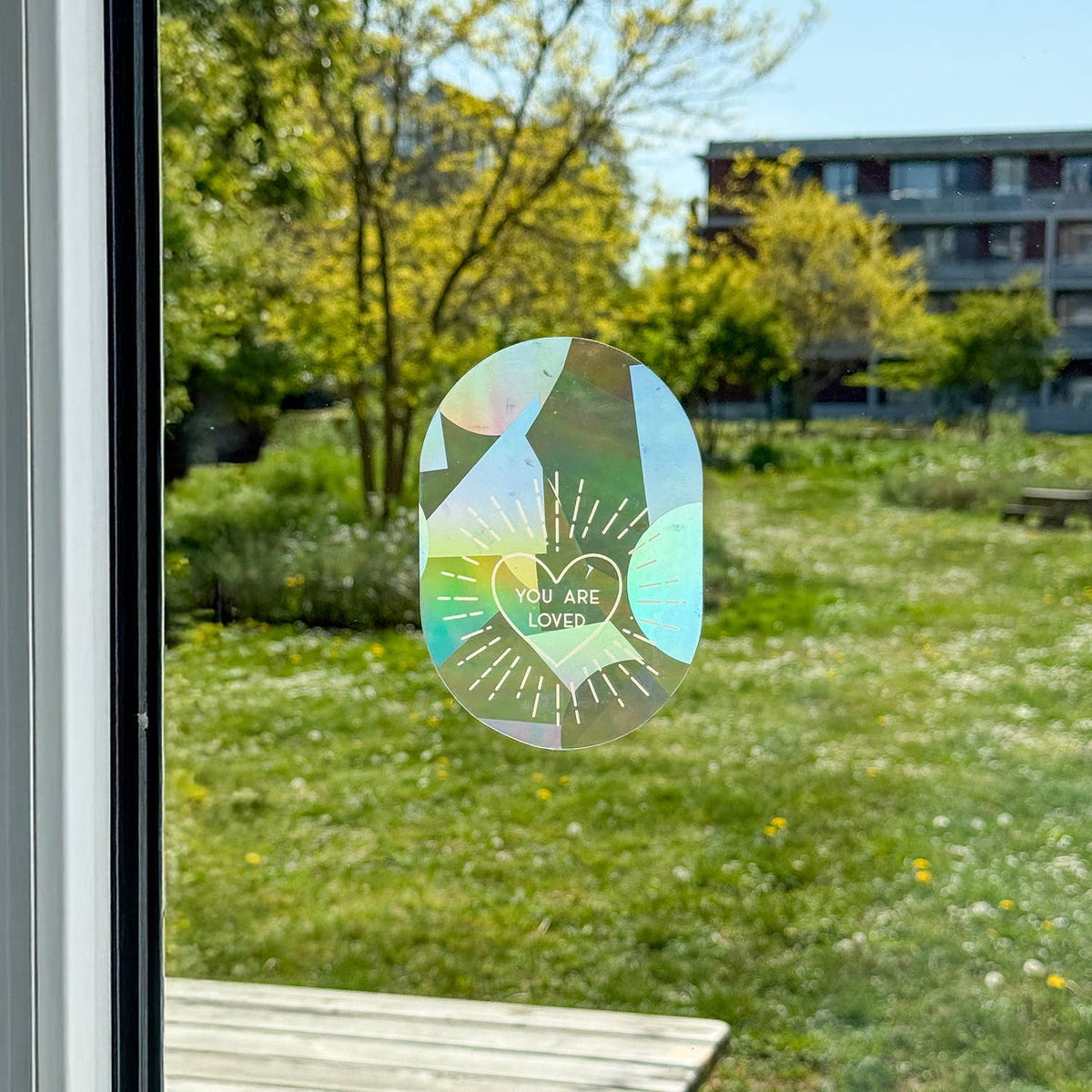 Fenstersticker mit Herzmotiv und „You Are Loved“ auf einer Scheibe mit Blick ins Grüne.