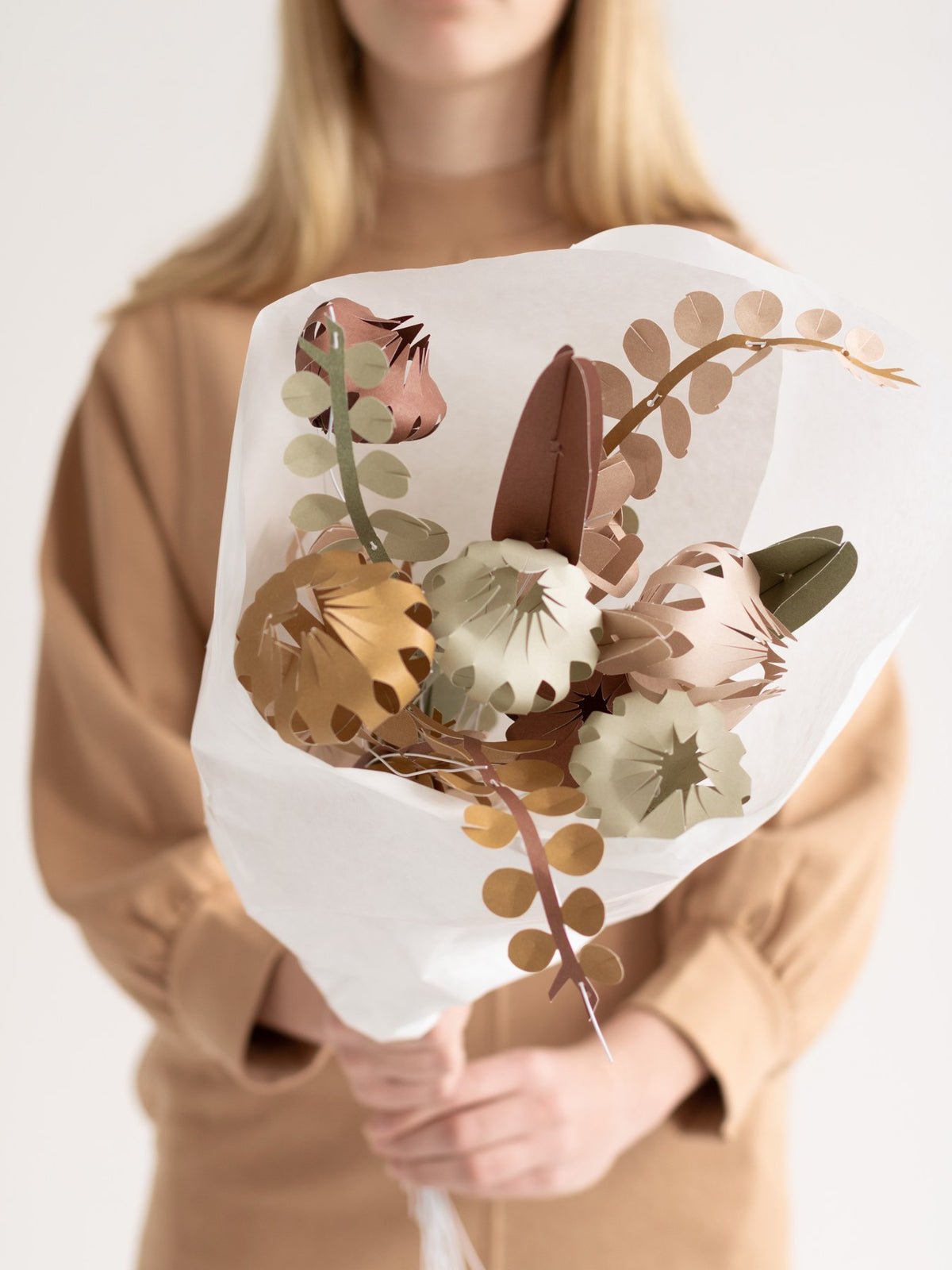 Person hält einen FIELD-Papierblumenstrauß in sanften Grün-, Rosé- und Brauntönen, eingeschlagen in weißes Papier.